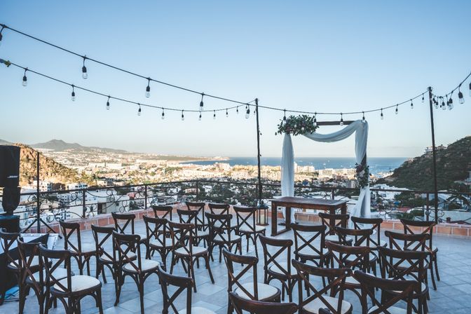 Rooftop terrace outdoor wedding setup with rows of wooden cross-back chairs, a draped floral arch, and hanging string lights overlooking a coastal town and blue ocean under a clear sky