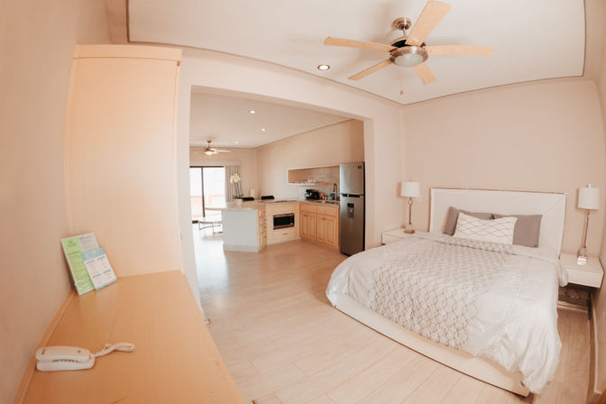 Sunlit open-plan studio apartment with a large bed, bedside lamps, ceiling fan, light wood floors, kitchen island, stainless refrigerator and sliding balcony doors.