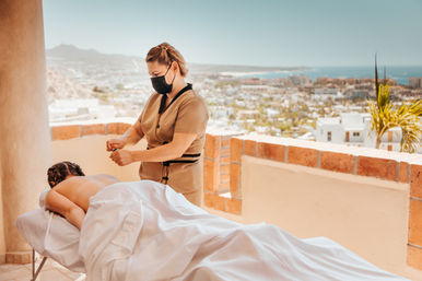 Sunlit rooftop massage: a masked therapist gives a relaxing treatment to a client on a table, overlooking a coastal cityscape and ocean from a terraced resort view.
