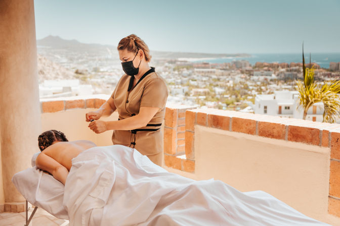 Sunlit rooftop massage: a masked therapist gives a relaxing treatment to a client on a table, overlooking a coastal cityscape and ocean from a terraced resort view.