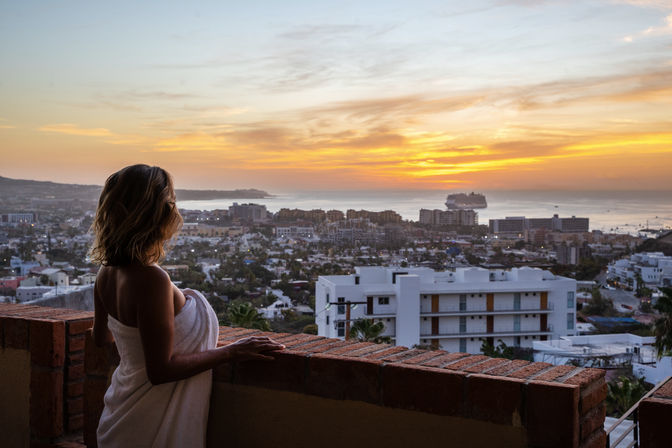 Woman wrapped in a towel leans on a brick balcony, gazing at a golden ocean sunset over a coastal city skyline with a cruise ship on the horizon.