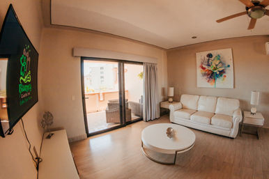 Bright modern condo living room with white leather sofa, round coffee table, colorful abstract wall art, sliding glass doors to a sunny balcony with wicker chairs and a ceiling fan