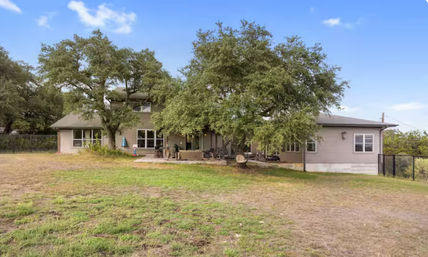 Ranch-style two-story house with a wide patio shaded by mature oak trees and a spacious grassy backyard under a clear blue sky.
