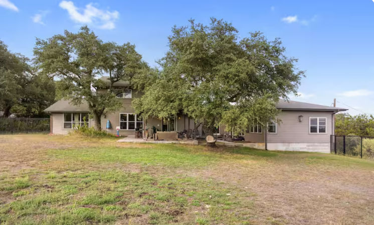 Ranch-style two-story house with a wide patio shaded by mature oak trees and a spacious grassy backyard under a clear blue sky.