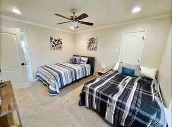 Cozy neutral guest bedroom with two striped-bedding beds, ceiling fan, beige carpet, wooden nightstand and lamp, and botanical wall art
