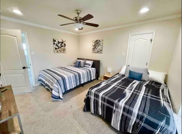 Cozy neutral guest bedroom with two striped-bedding beds, ceiling fan, beige carpet, wooden nightstand and lamp, and botanical wall art