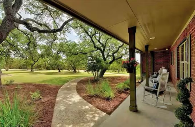 Brick front porch with rocking chairs and hanging flowers, curved concrete walkway leading through landscaped beds to an oak‑shaded lawn