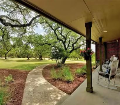 Brick front porch with rocking chairs and hanging flowers, curved concrete walkway leading through landscaped beds to an oak‑shaded lawn