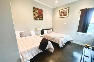 Bright guest bedroom with two full beds in neutral bedding, bedside table with lamp, framed wall art, dark polished floor and a curtained window letting in natural light