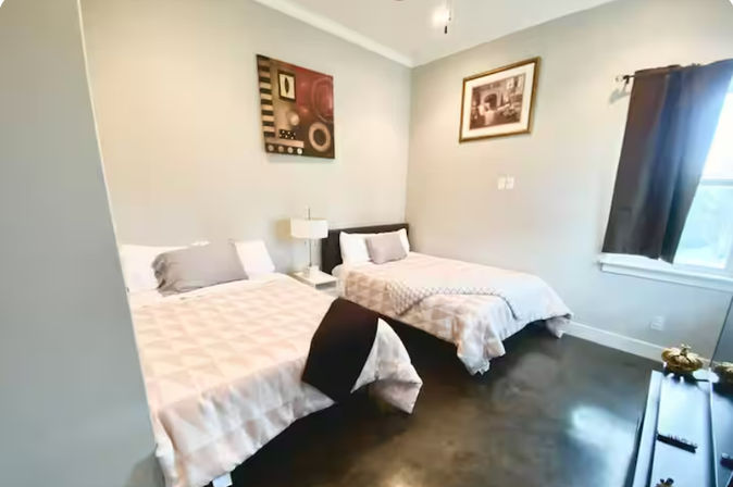 Bright guest bedroom with two full beds in neutral bedding, bedside table with lamp, framed wall art, dark polished floor and a curtained window letting in natural light
