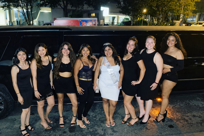 Nine women in party attire — eight in black outfits and one in white wearing a sash and tiara — smiling and posing in front of a black stretch limousine on a city street at night.
