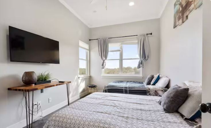 Bright, cozy modern guest bedroom with wall-mounted flat-screen TV, slim wooden console table, two beds with gray bedding, and a large sunlit window with tied-back curtains.