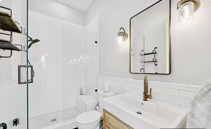 Bright modern white bathroom with glass walk-in shower, white subway tile, brass faucet and hardware, black-framed rectangular mirror, wood vanity drawer and wall sconces