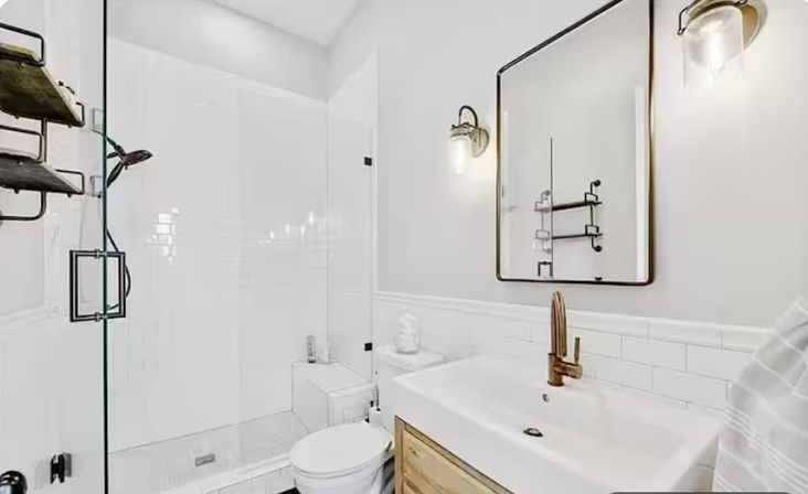 Bright modern white bathroom with glass walk-in shower, white subway tile, brass faucet and hardware, black-framed rectangular mirror, wood vanity drawer and wall sconces