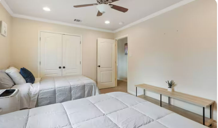 Bright neutral guest bedroom with two twin beds dressed in light-gray comforters, ceiling fan, double closet doors, and a minimalist wooden bench with a small potted plant.
