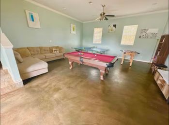 Bright family game room with a red-felt pool table centered on polished concrete floors, beige sectional sofa, ping-pong and foosball tables, light blue walls, ceiling fan and two windows letting in natural light.