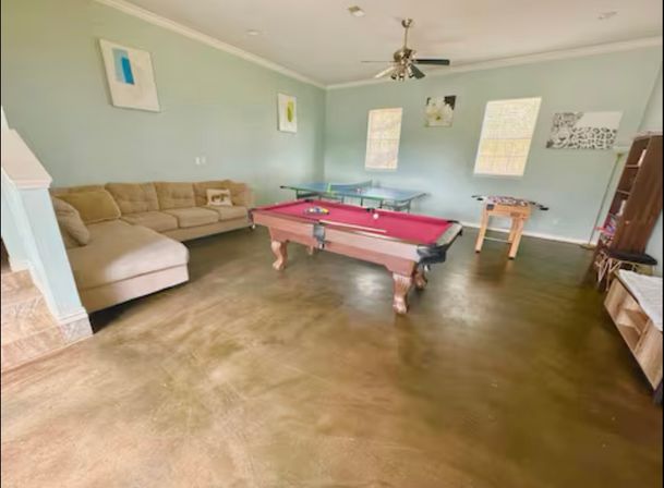 Bright family game room with a red-felt pool table centered on polished concrete floors, beige sectional sofa, ping-pong and foosball tables, light blue walls, ceiling fan and two windows letting in natural light.
