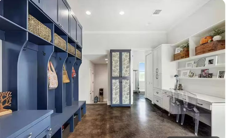 Modern residential mudroom and home office with navy built-in lockers and bench cubbies, woven baskets and hooks for bags, patterned tall cabinet, white built-in desk with clear acrylic chairs, decorative shelves and polished concrete floor.