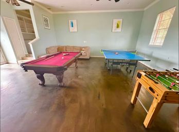 Home game room ready for game night with a pink-felt pool table, blue ping-pong table and wooden foosball table, beige sofa, mint-green walls and framed abstract art.