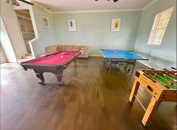 Home game room ready for game night with a pink-felt pool table, blue ping-pong table and wooden foosball table, beige sofa, mint-green walls and framed abstract art.