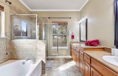 Bright, spa-like master bathroom with long wood double vanity and granite counters, glass-enclosed walk-in shower, soaking tub, tiled floors and window view of trees.