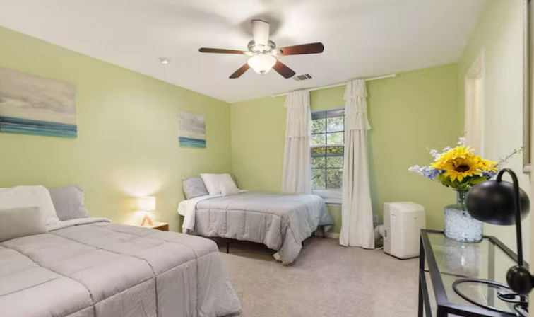 Bright, cheerful bedroom with two twin beds in gray bedding, light green walls, ceiling fan, window with ruffled white curtains, bedside lamp and glass desk with sunflowers.