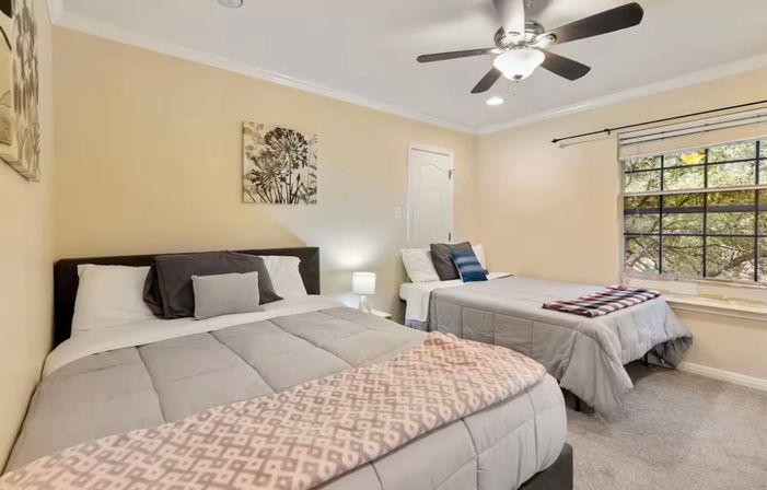 Cozy guest bedroom with two beds in gray comforters, beige walls, floral wall art, bedside lamp, ceiling fan and a large window with a tree-lined view