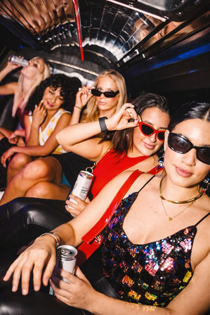 Five friends enjoying a night out inside a party limo, wearing sunglasses and colorful outfits, holding canned drinks and posing for the camera.