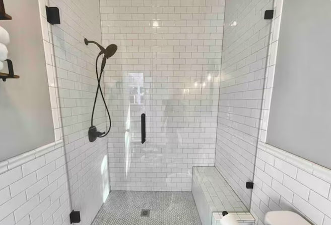 Bright modern bathroom walk-in shower with white subway tile walls, frameless glass enclosure, matte black handheld shower and fixtures, tiled bench and patterned mosaic floor.