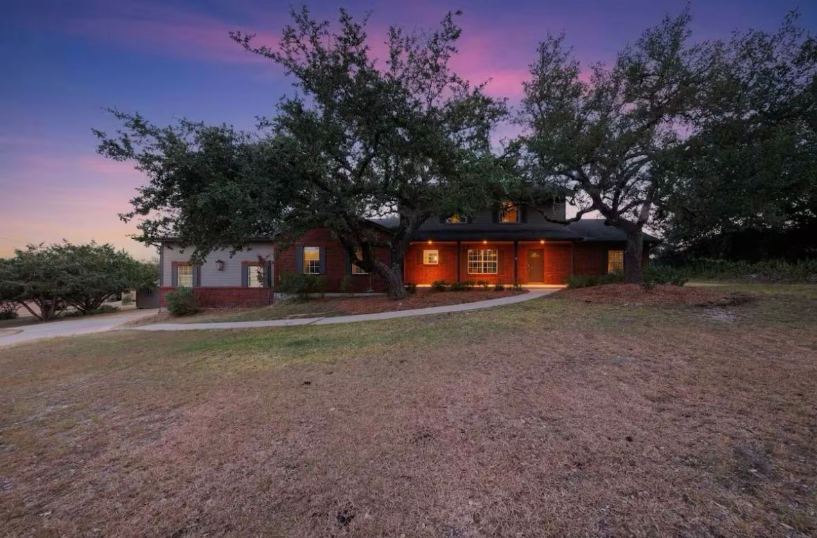 Ranch-style brick home at dusk with glowing front porch lights, large oak trees, curved driveway and dry lawn beneath a purple-pink sky