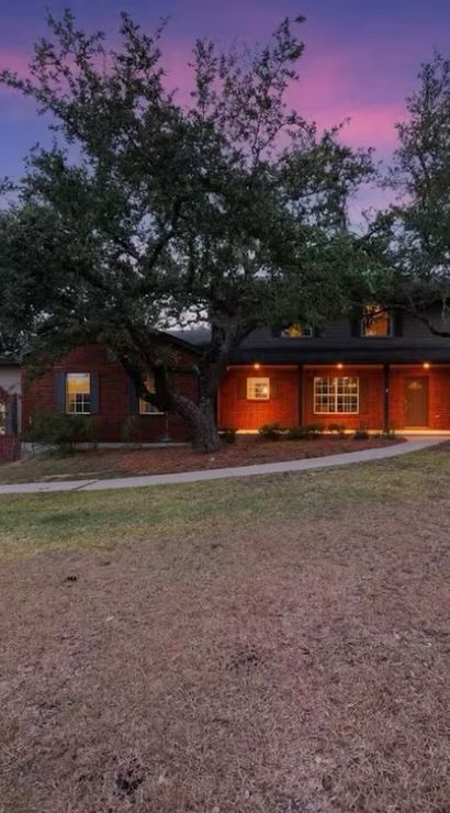 Ranch-style brick home at dusk with glowing front porch lights, large oak trees, curved driveway and dry lawn beneath a purple-pink sky