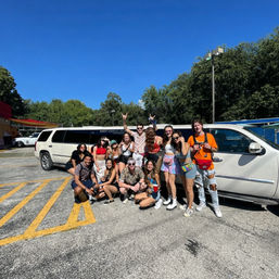 Cheerful group of young adults in colorful festival outfits posing in front of a white stretch SUV limousine in a sunny parking lot under a bright blue sky with trees in the background.