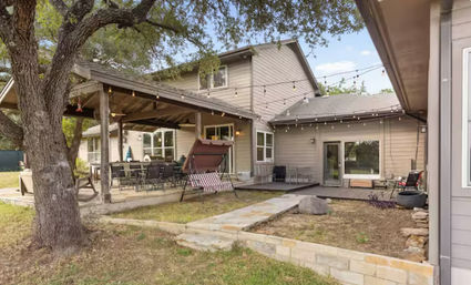 Cozy suburban backyard patio of a two-story home with a covered porch and string lights, outdoor dining table, striped swing, stone walkway and large oak tree.