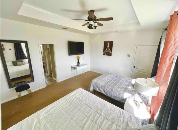 Cozy sunlit bedroom in a suburban home with two beds, tray ceiling and ceiling fan, wall-mounted TV, full-length mirror, bedside lamp, coral curtains and tan carpet.