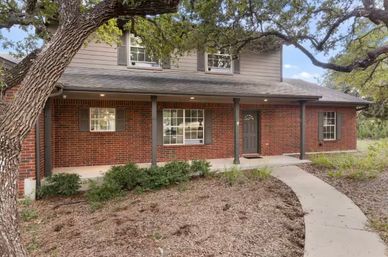 Charming two-story red brick house with covered front porch, gray shutters and door, large oak tree overhanging a curved concrete walkway and low shrubs