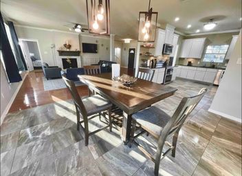 Sunlit open-concept kitchen and dining area with a wooden table and four chairs, pendant lights, gray stone-look tile floor, white cabinets and stainless appliances, and an adjoining living room with fireplace and wall-mounted TV.