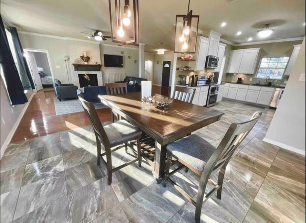 Sunlit open-concept kitchen and dining area with a wooden table and four chairs, pendant lights, gray stone-look tile floor, white cabinets and stainless appliances, and an adjoining living room with fireplace and wall-mounted TV.