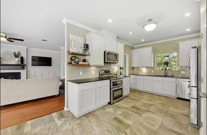 Bright open-concept kitchen with white cabinets, stainless-steel appliances (double oven and microwave), black countertops, large beige stone-look tile floor, sink under a window, and view into a cozy living room with fireplace and wall-mounted TV.
