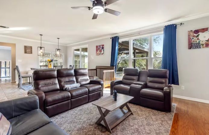 Open-plan family living room with dark brown leather reclining sofa and two recliners, wooden coffee table on a patterned rug, baby crib by large windows with navy curtains, dining area with pendant lights in the background and a ceiling fan overhead.