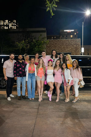 Smiling group of friends in pink party outfits posing at night beside a black stretch limousine on a downtown street, city buildings and a lit hotel sign in the background.