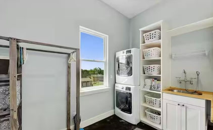 Bright laundry room with stacked washer and dryer, built-in shelving of laundry baskets, utility sink and a window with neighborhood view