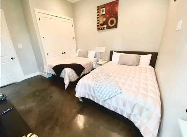 Cozy modern guest bedroom with two beds in light geometric bedding, shared white nightstand and lamp, abstract wall art, white closet doors and polished dark concrete floor.