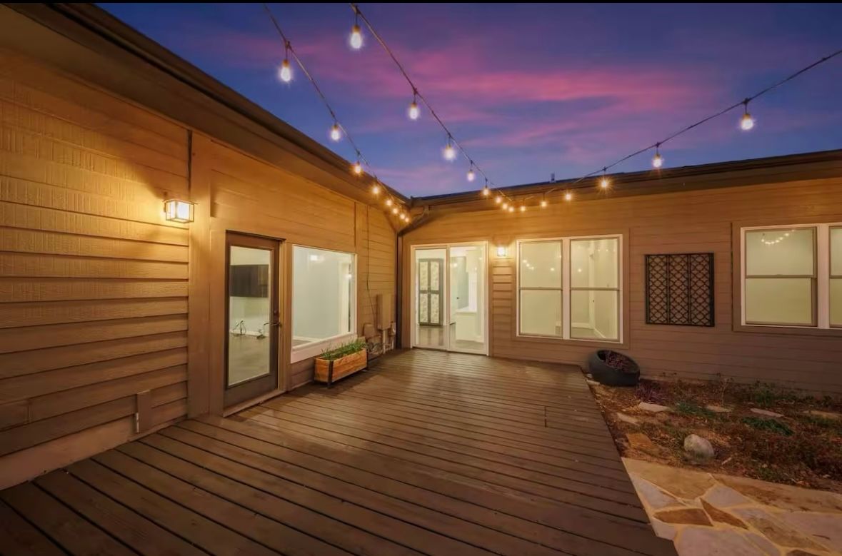Cozy evening backyard patio with wood deck, hanging string lights and warm wall sconces, planter box and glass doors opening into a modern home under a purple twilight sky.