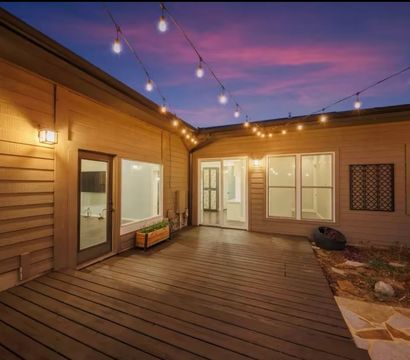 Cozy evening backyard patio with wood deck, hanging string lights and warm wall sconces, planter box and glass doors opening into a modern home under a purple twilight sky.