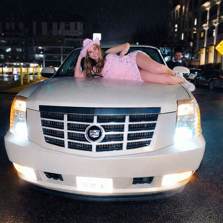 Smiling woman in a pink sequin dress and cowboy hat lounging on the hood of a white Cadillac SUV with headlights on, parked on a wet city street at night.