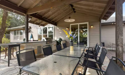 Covered backyard patio with vaulted wood ceiling, long glass-top dining tables and black mesh chairs, stone outdoor kitchen with built-in grill, vase of flowers, and sliding glass door to the house.