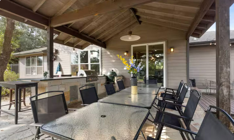 Covered backyard patio with vaulted wood ceiling, long glass-top dining tables and black mesh chairs, stone outdoor kitchen with built-in grill, vase of flowers, and sliding glass door to the house.