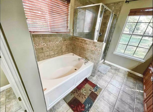 Sunny master bathroom with a white corner soaking tub, glass-enclosed tile shower, beige mosaic wall tile, large window with wooden blinds, tiled floor and colorful bath mat