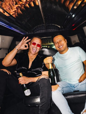 Two friends partying in a limousine at night, smiling and holding champagne bottles — one wearing red sunglasses and flashing an OK sign.