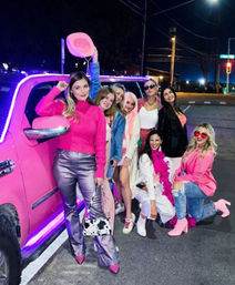 Eight friends in pink-themed outfits posing by a pink limousine with purple LED trim on a downtown street at night, one holding a cowboy hat and another wearing a feather boa and heart-shaped sunglasses.